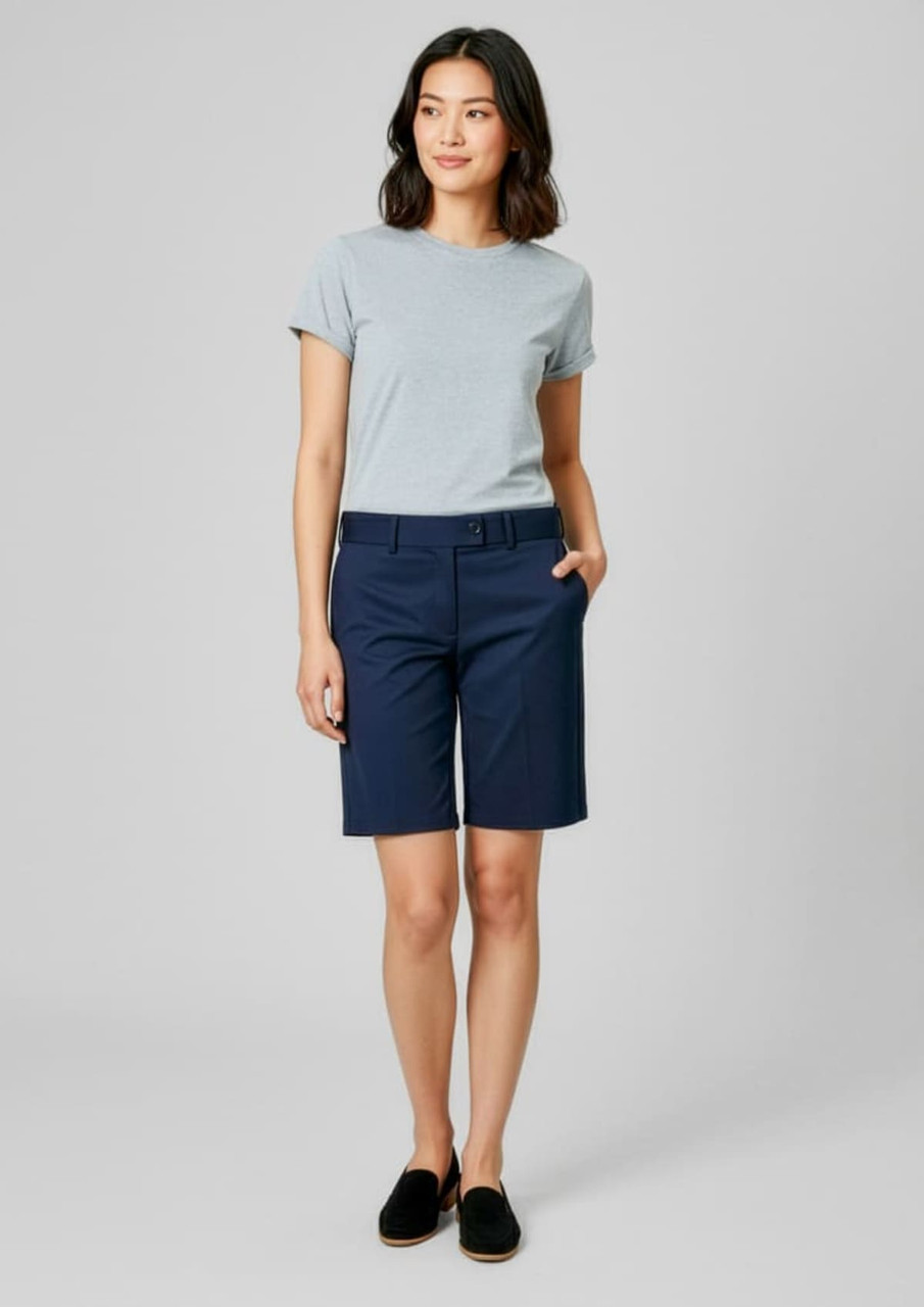 A woman stands wearing navy dress shorts and a light grey T-shirt, with her hands in the pockets. The image features a logo.
