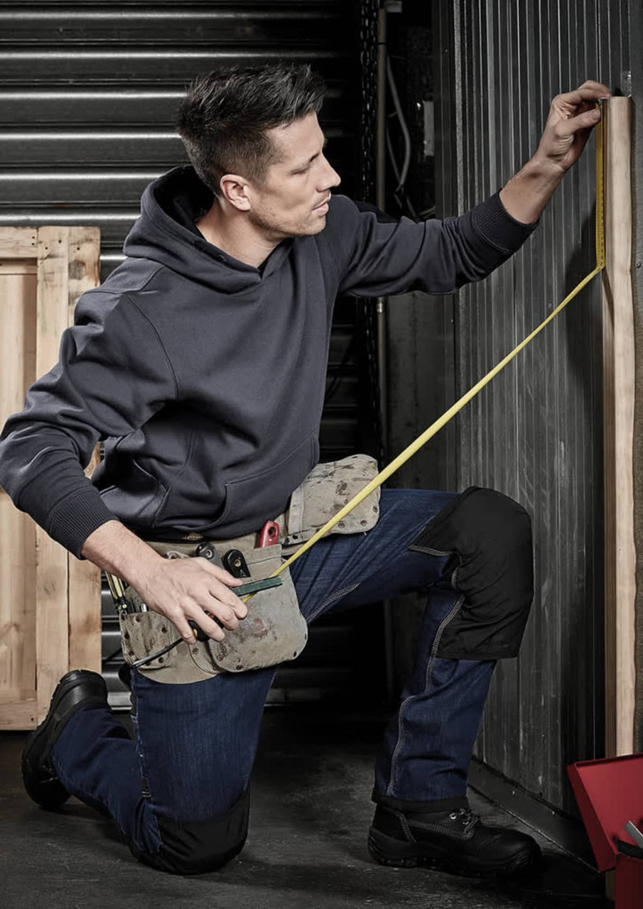 A man in a grey hoodie and dark trousers measures a wooden plank while kneeling, wearing a tool belt.