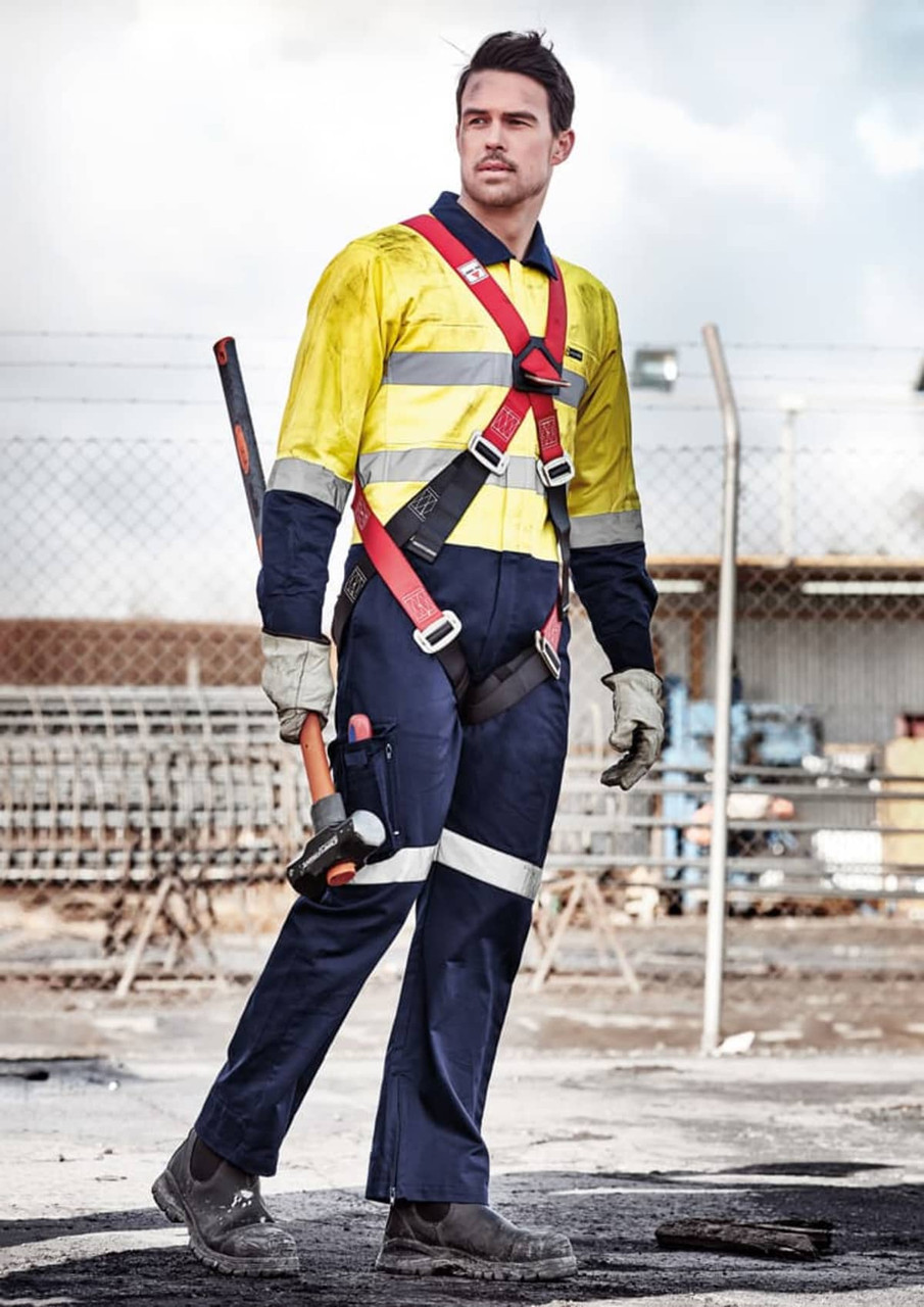 A man wears a yellow and navy rugged cooling overall with safety features. He holds a tool and stands in an industrial setting.
