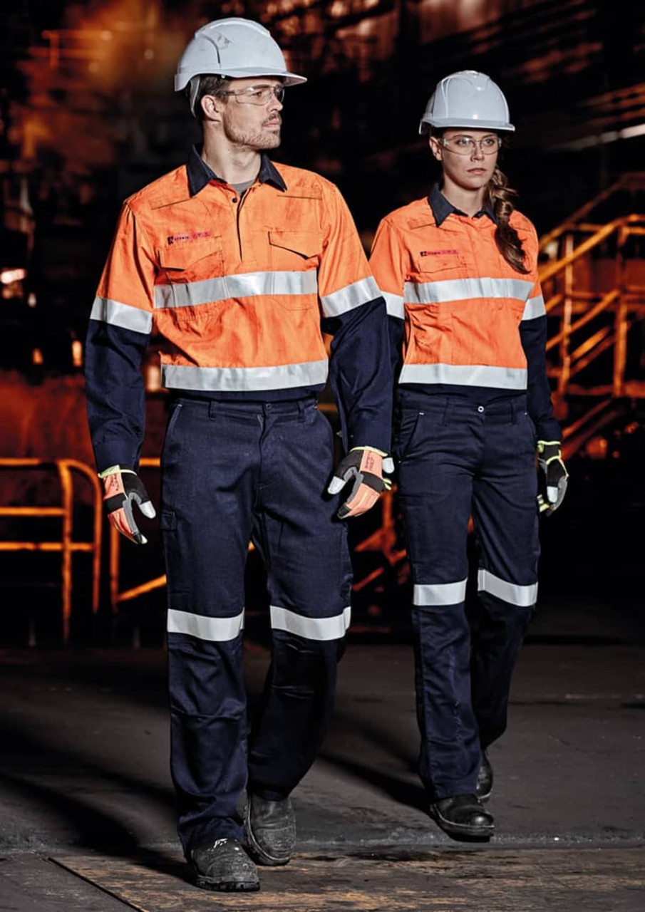 Two individuals wearing orange and navy safety shirts and pants, equipped with hard hats and gloves, in an industrial setting.