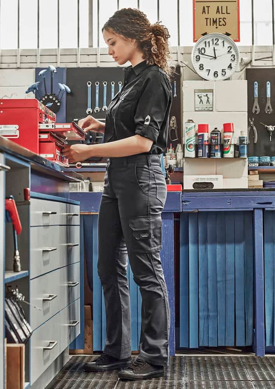 A woman in black rugged cargo pants stands at a workshop bench, engaging with tools and equipment. The pants feature pockets and a logo.