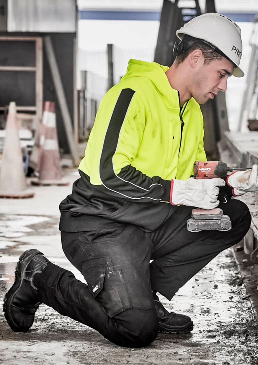 A unisex hi vis full zip hoodie in bright yellow, worn by a person kneeling and working on a construction site.