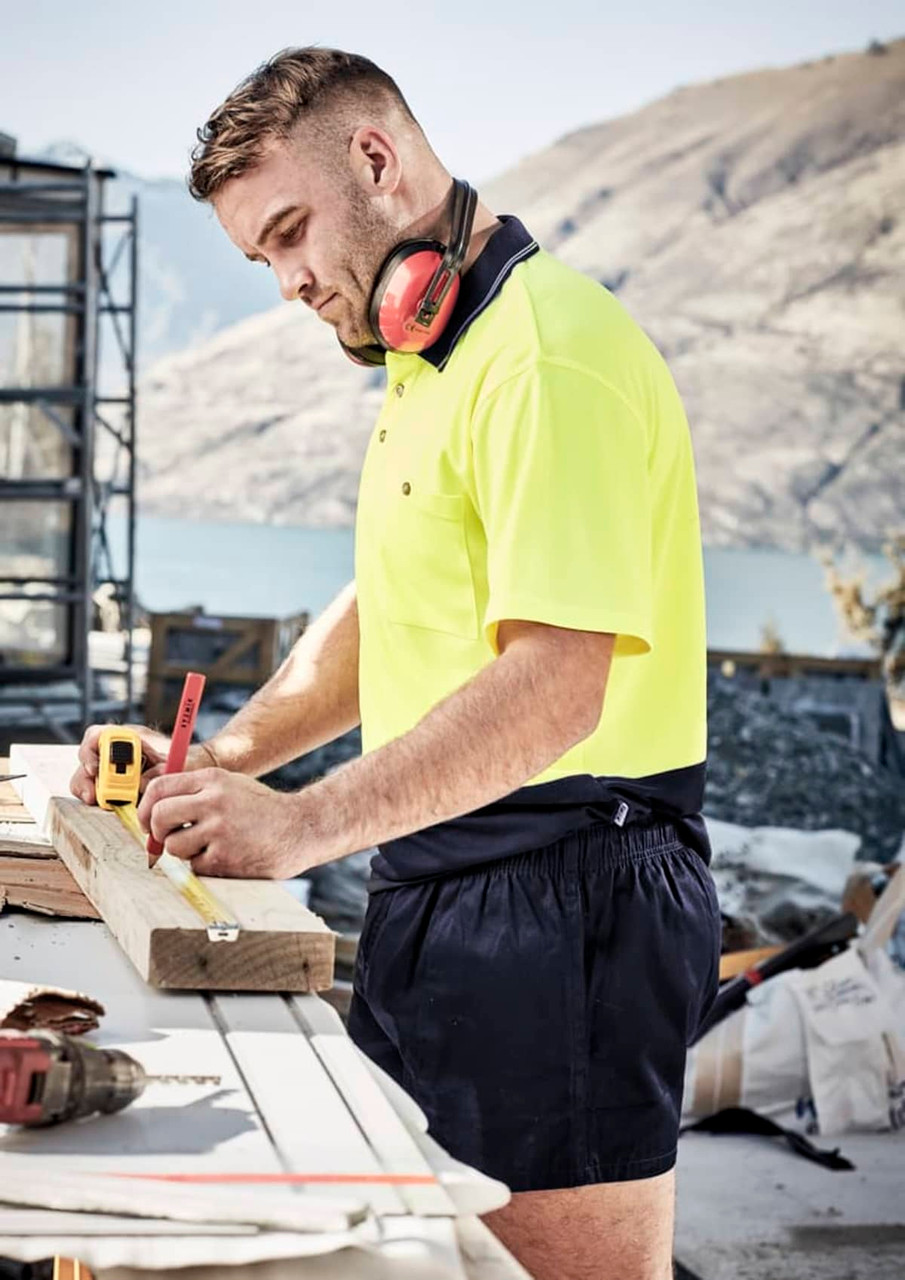 A man in a high-visibility yellow shirt and navy rugby shorts measures a wooden plank at a construction site.