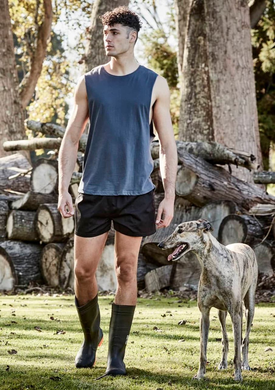 A man in a sleeveless top and black rugby shorts walks beside a greyhound in a natural setting.