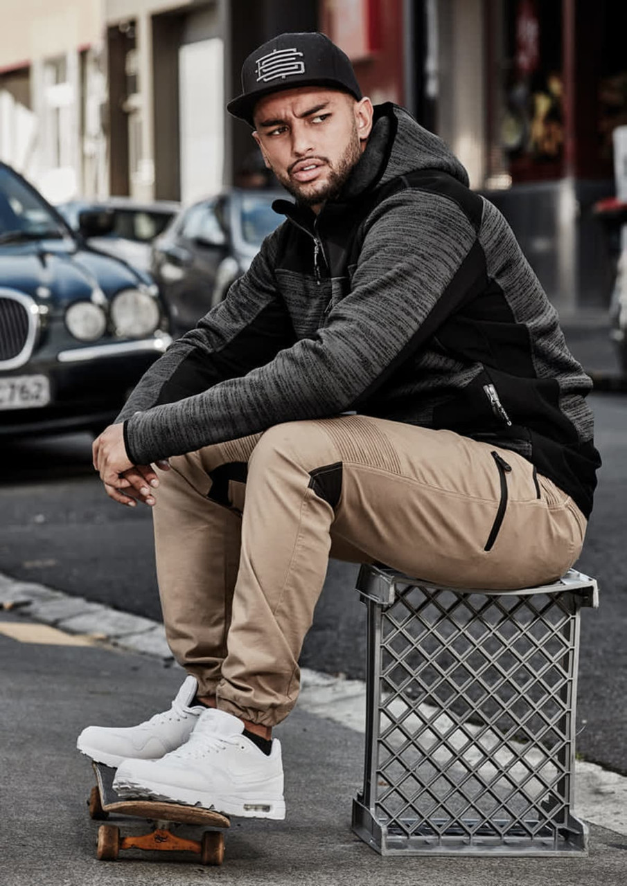 A model wears a black and grey hoodie with reinforced details and khaki pants, sitting on a crate in an urban setting.