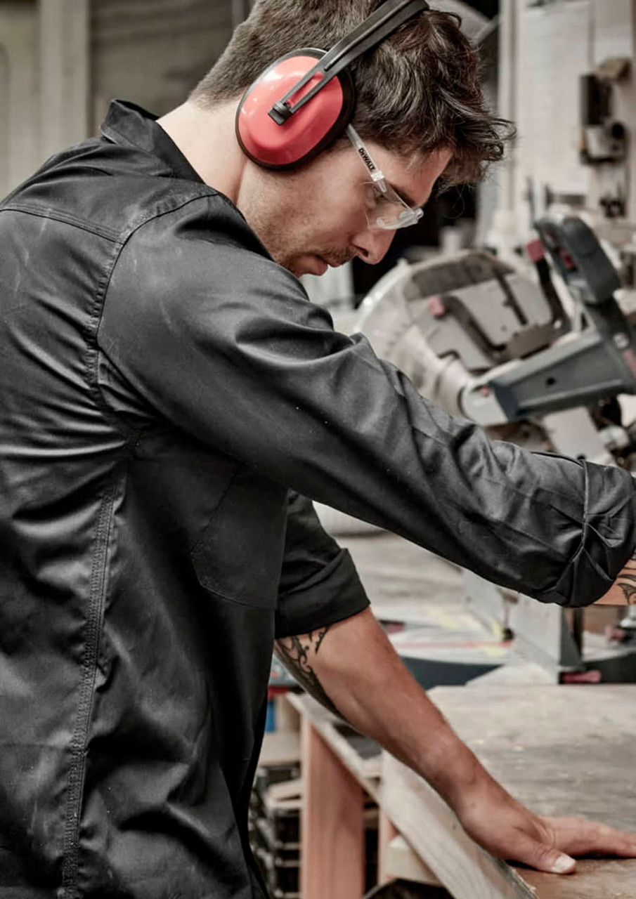 A man wearing a black long sleeve shirt, safety glasses, and earmuffs, working at a bench with machinery.