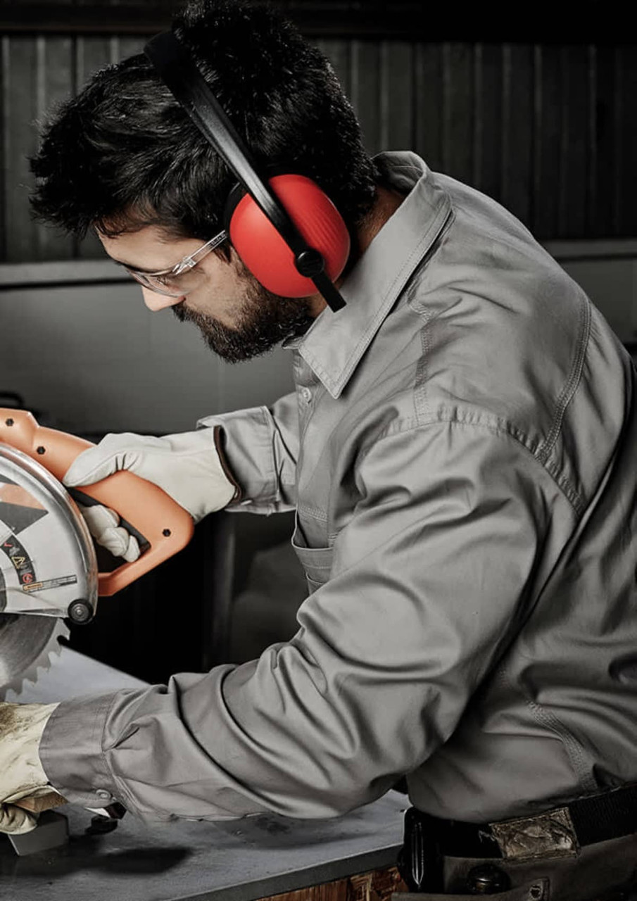 A man wearing a grey long sleeve shirt and safety gear operates a power saw in a work environment.