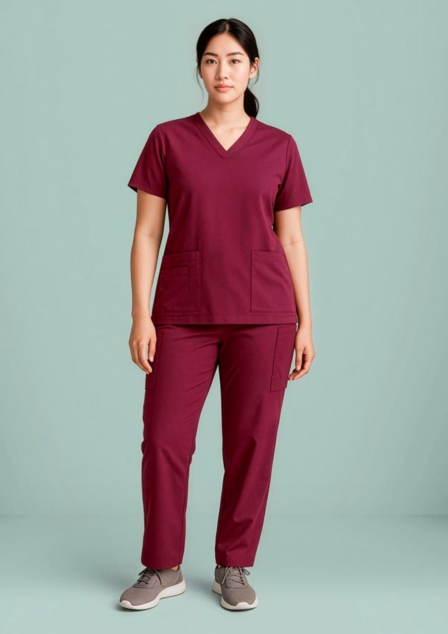 A woman stands wearing burgundy straight leg scrub pants and a matching short-sleeve top. The outfit has two pockets.