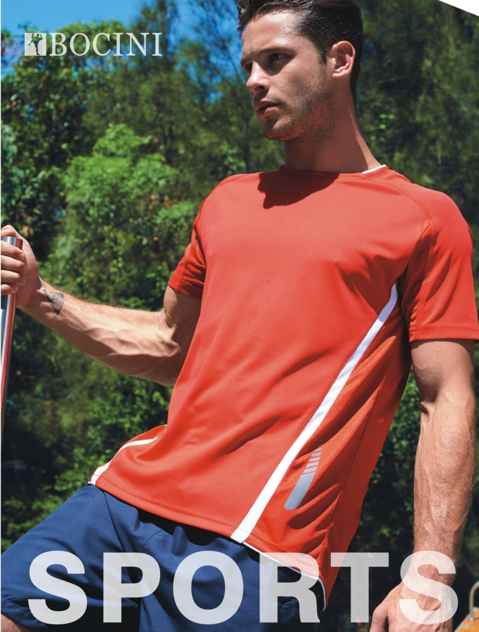 A man models an orange sports tee with grey accents, paired with navy shorts, set against a green outdoor backdrop.