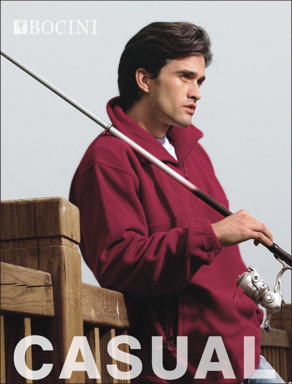 A man wearing a maroon polar fleece jumper stands by a wooden railing, holding a fishing rod.