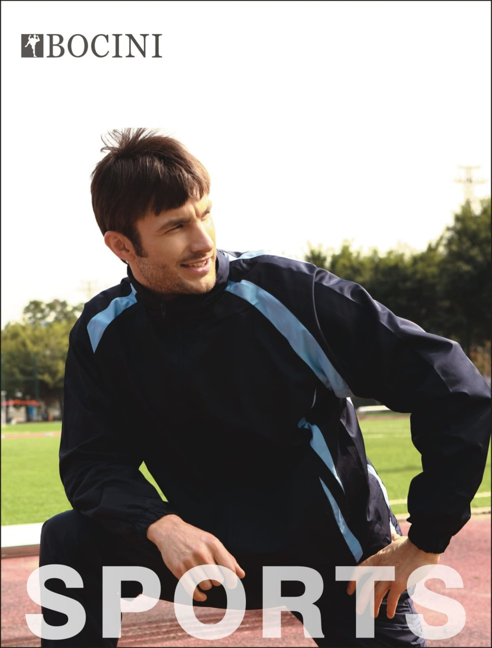 A man wearing navy blue track pants with light blue accents, posing on a sports field. The outfit includes a matching jacket and has a logo.