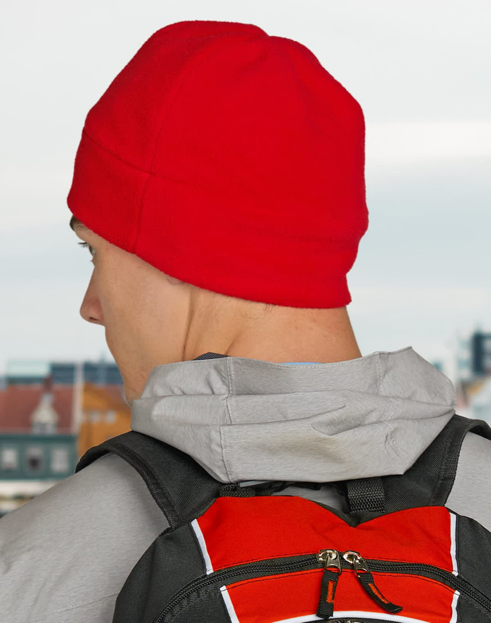 A red fleece beanie worn by a person with a grey hoodie and a black backpack. The background features a cityscape.