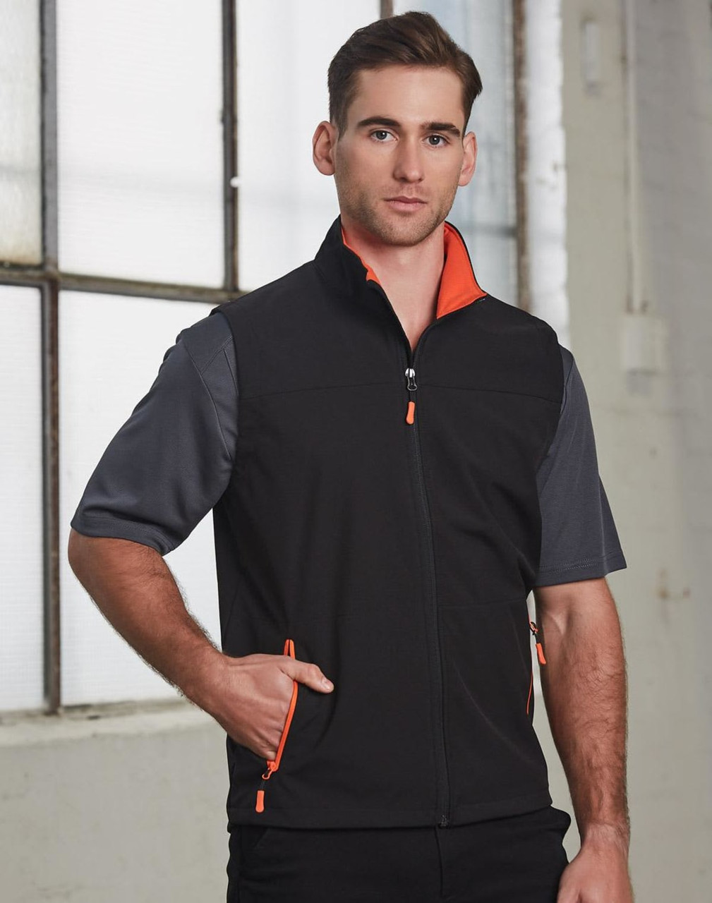 A men's softshell vest in black with orange accents, featuring a zip front and side pockets. It has a logo on it.