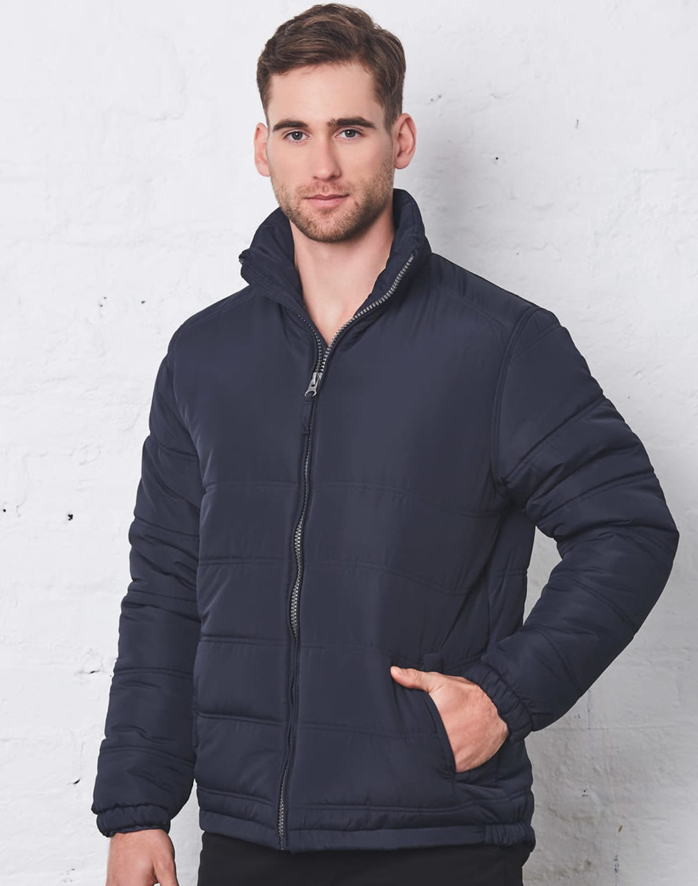 A navy heavy quilted puffer jacket with a zippered front and high collar, modelled by a man against a white wall.