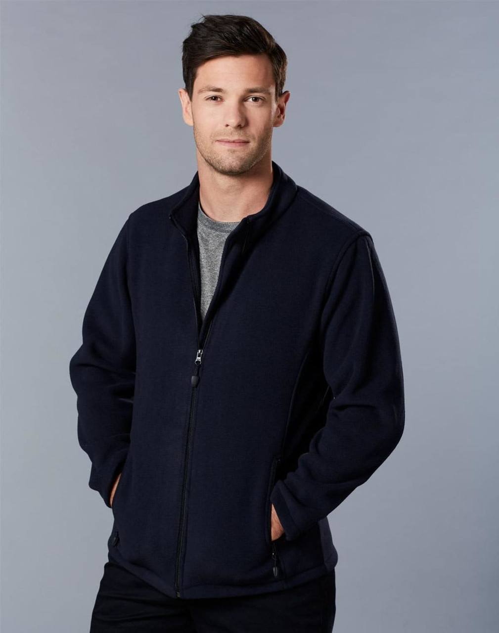 A man in a navy polar fleece jacket with a full zip, standing with hands in pockets against a grey background.