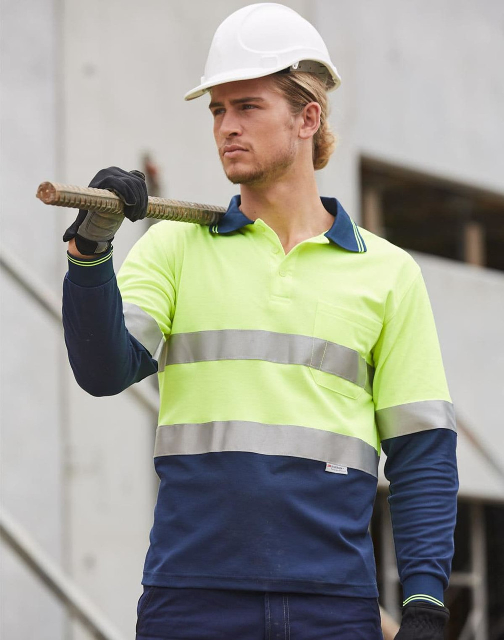 A long-sleeve work polo shirt in high-vis yellow and navy, featuring reflective tape and a pocket. A person wears a hard hat and holds a rod.