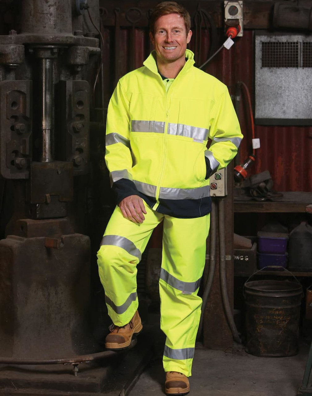 A man stands in a workshop wearing a high-visibility yellow safety pant and jacket with reflective 3M tapes.