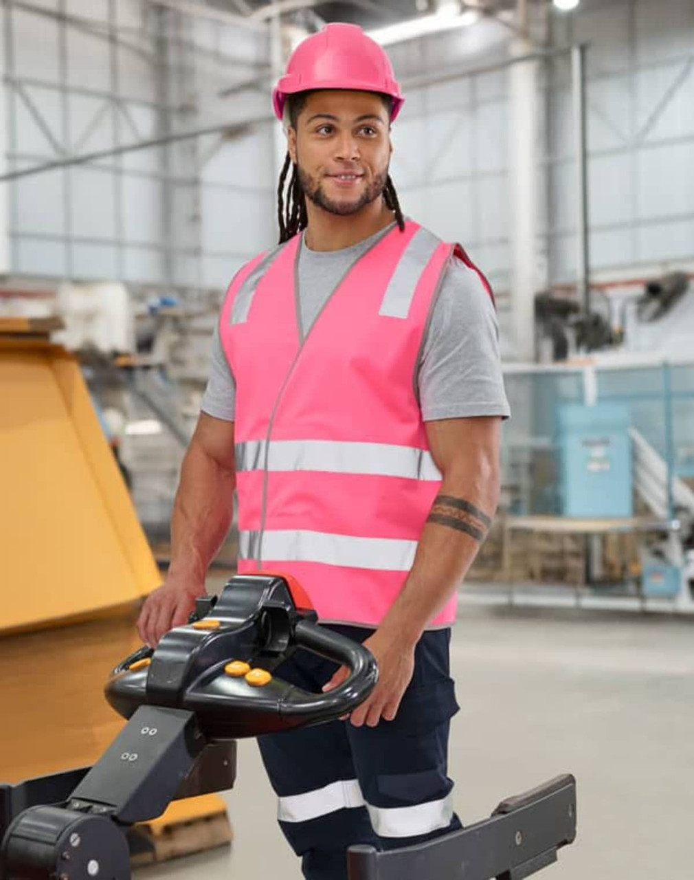A pink hi-vis safety vest with shoulder reflective tapes, worn by a person in a warehouse setting, featuring a logo.