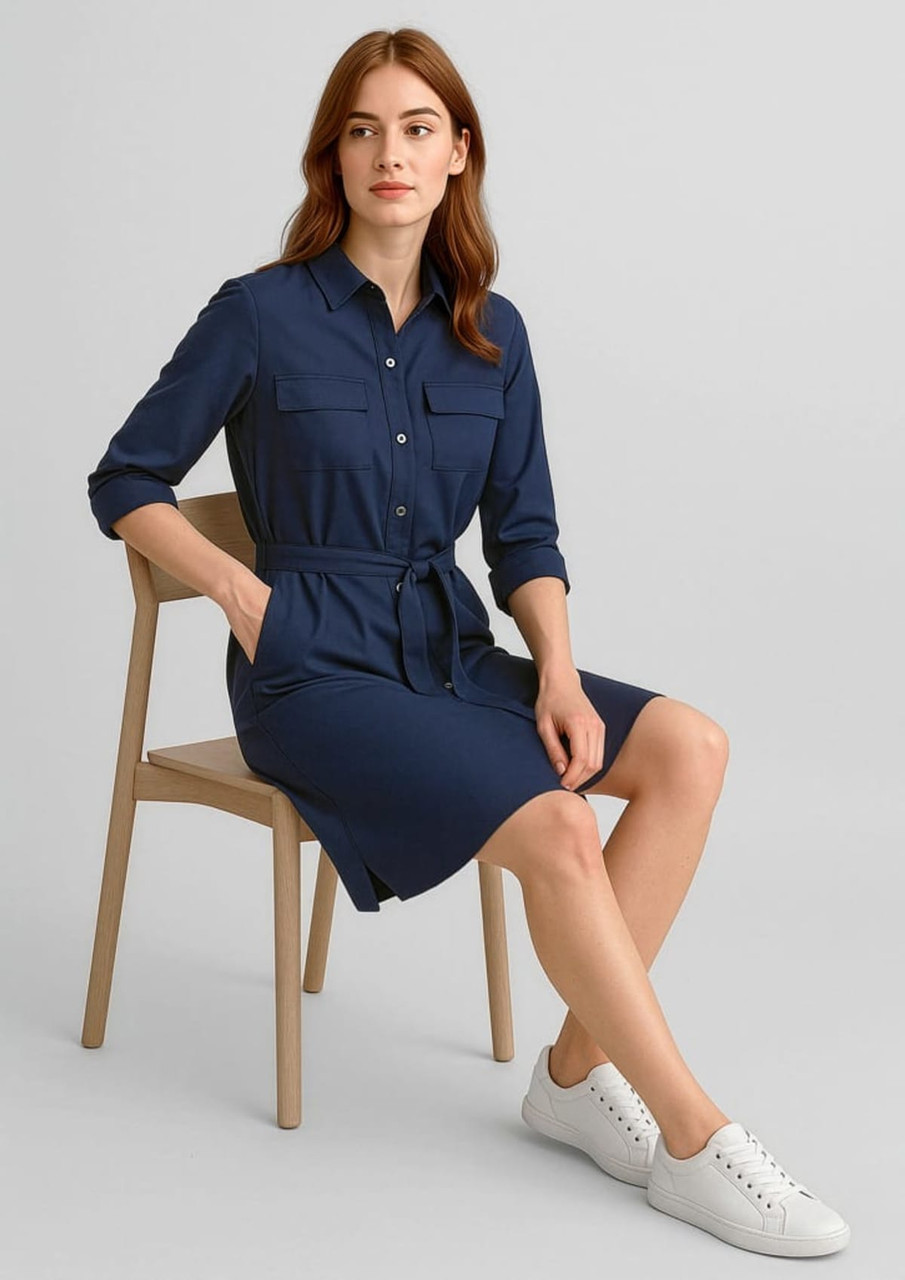 A navy blue dress with two pockets, roll-up sleeves, a belt, and a model seated on a wooden chair.