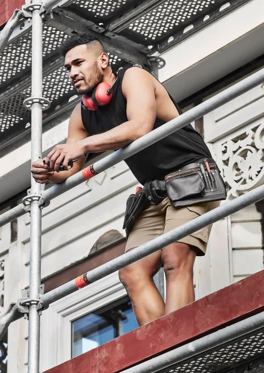 A man wearing a black sleeveless tee, shorts, and a tool belt stands on scaffolding, with headphones around his neck.