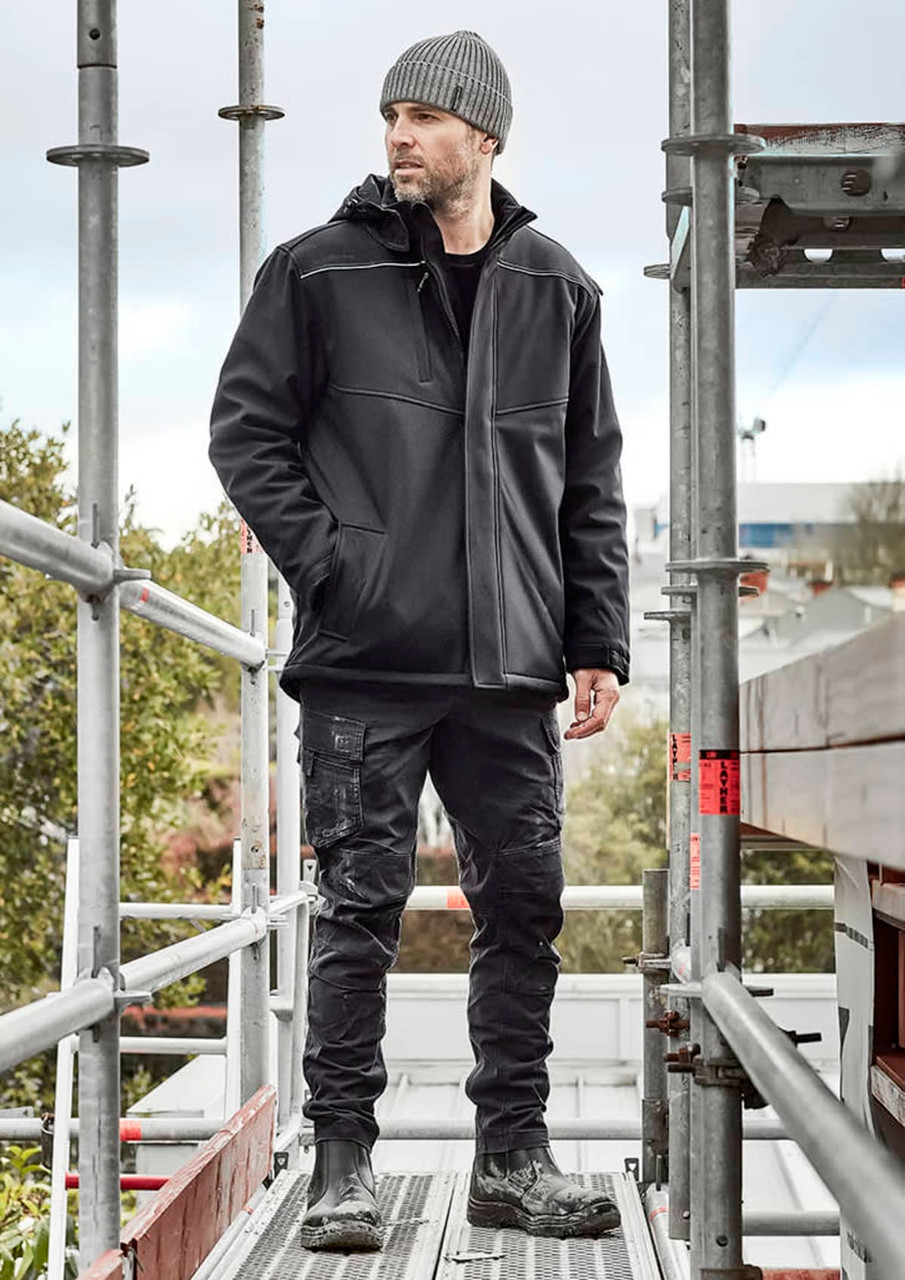 A man wearing a black unisex softshell jacket stands on scaffolding, paired with black trousers and boots.