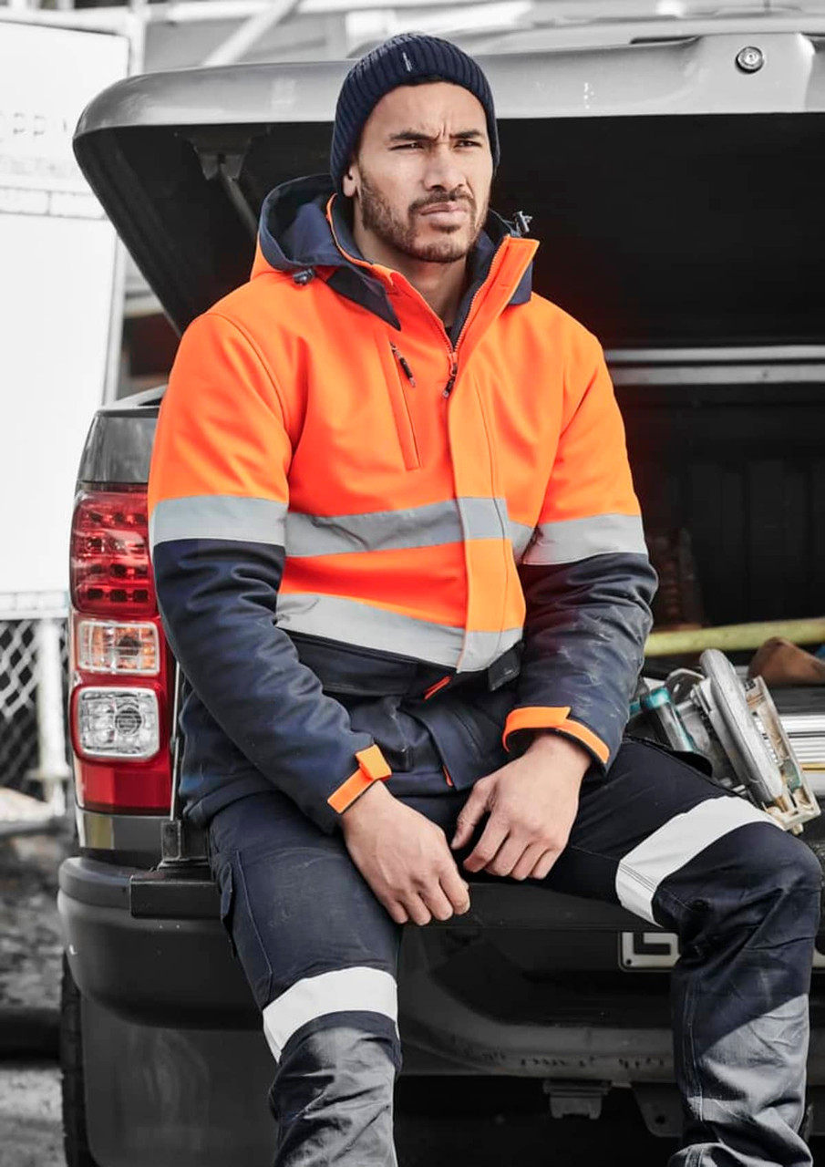A man wearing an orange and navy unisex hi-vis softshell jacket sits on the back of a vehicle.