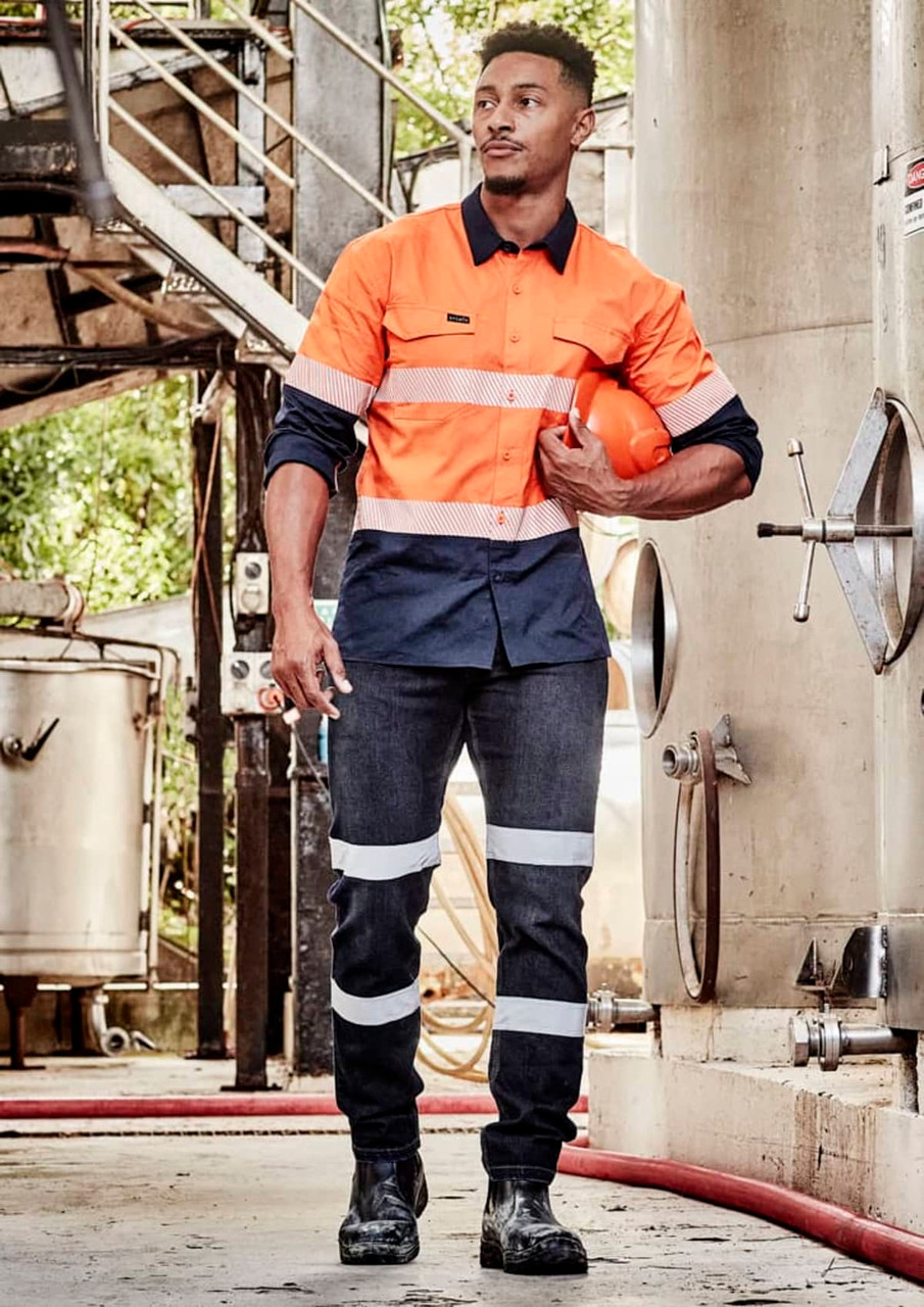 A worker in an orange and navy long sleeve shirt with segmented tape, holding a helmet in an industrial setting.