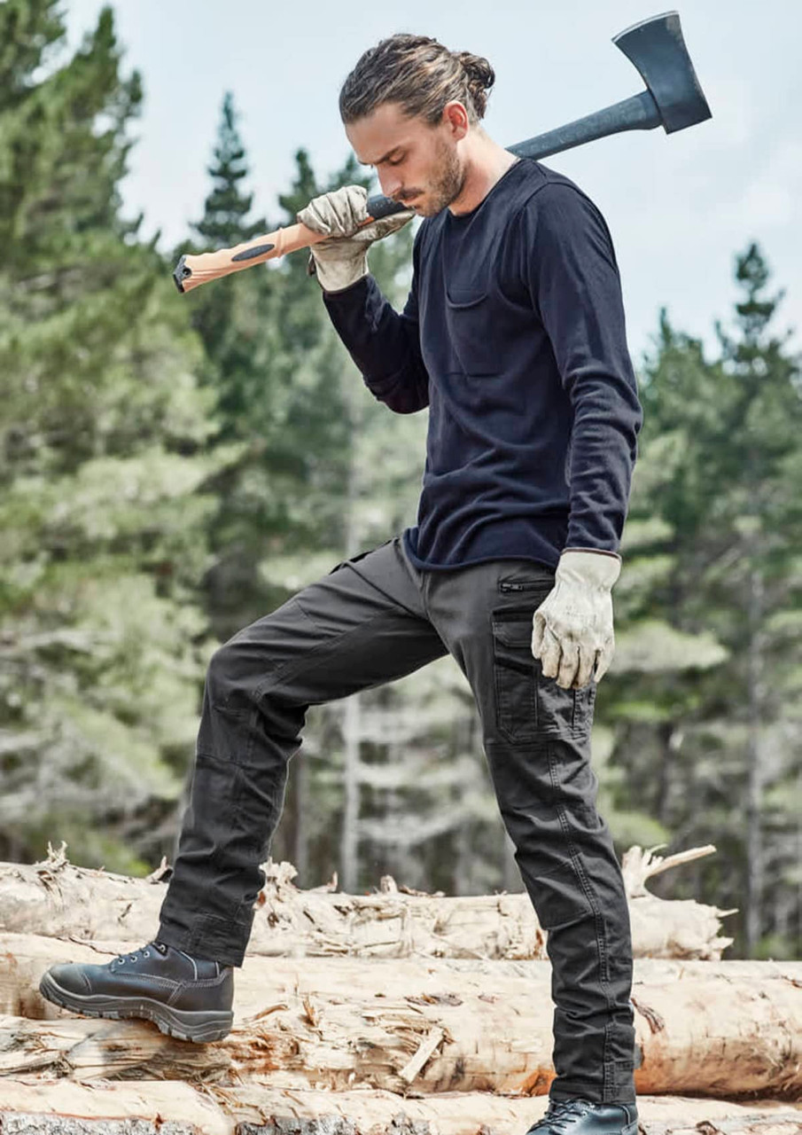 A man in black work pants and a long-sleeve top, holding an axe while standing on logs in a forested area.