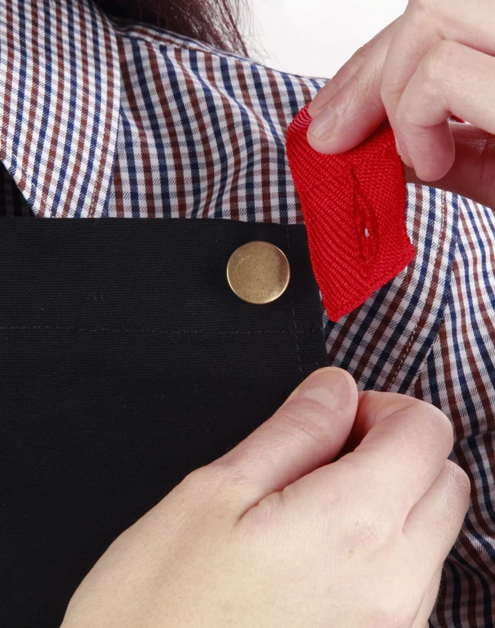 A black canvas bib apron is being adjusted, showcasing a button detail and a red fabric tag.