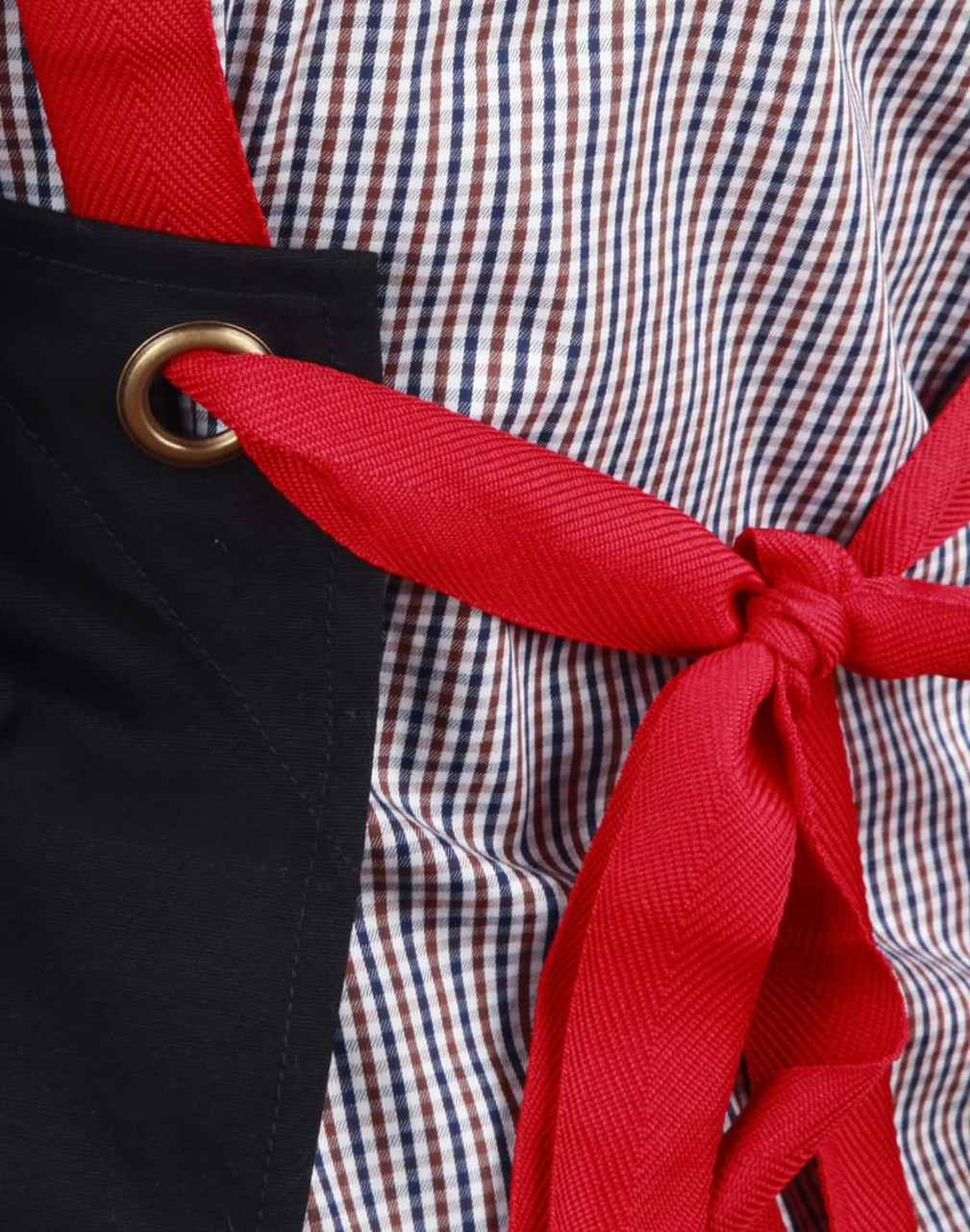 A black canvas bib apron with a red tie, featuring a grommet detail and a checkered fabric background.
