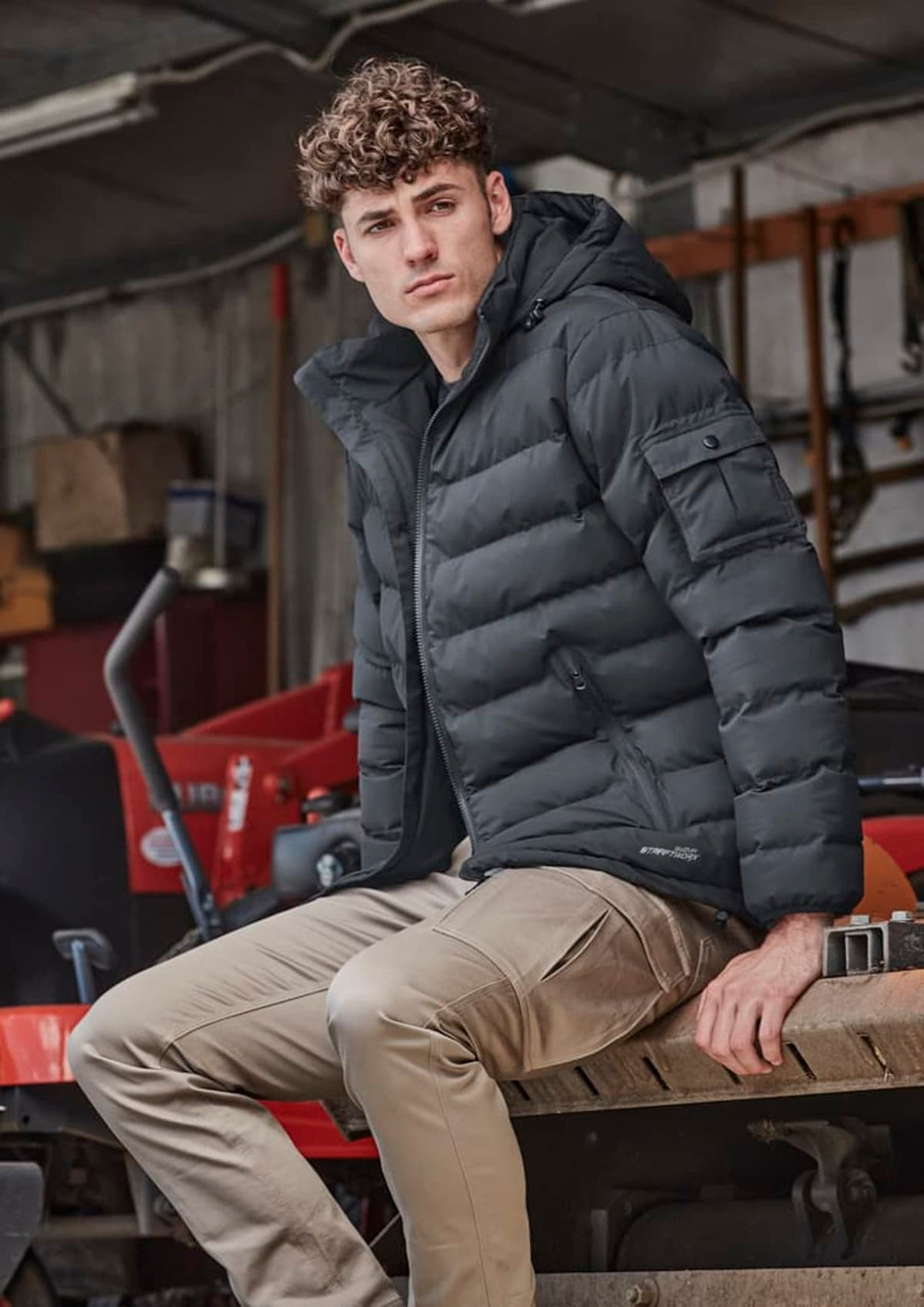 A unisex hooded puffer jacket in black, featuring a fitted design and a logo, worn by a model sitting on machinery.