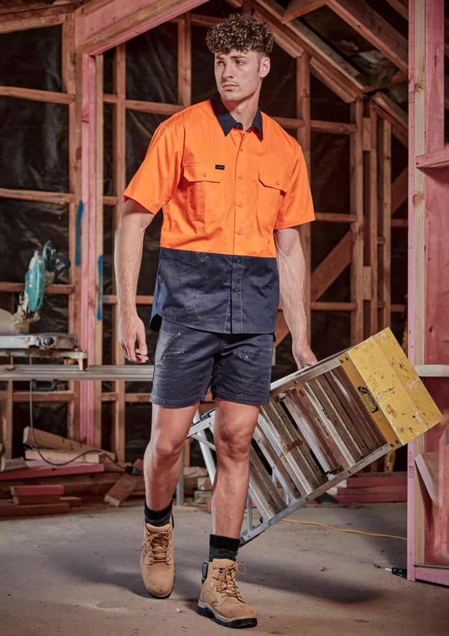 A man wearing an orange and navy work shirt and navy shorts carries a ladder in a construction setting.