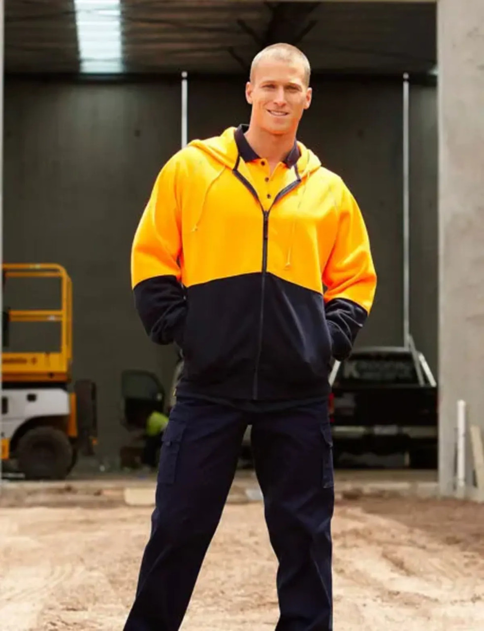 A man wearing a high-visibility orange and navy full zip fleece hoodie, posed in an industrial setting.