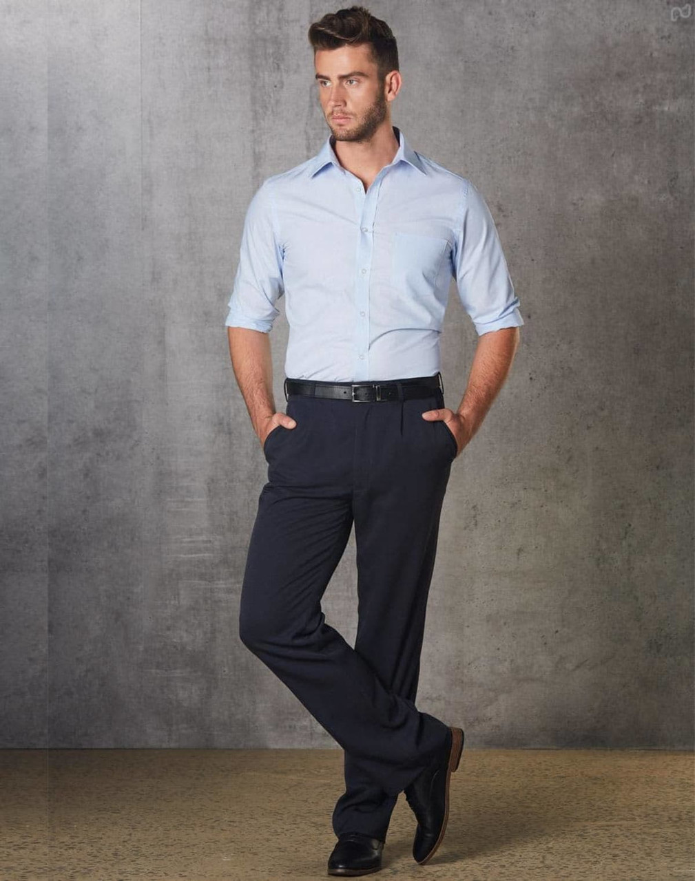 A man in a light blue shirt and black permanent press pants stands with hands in pockets against a grey backdrop.
