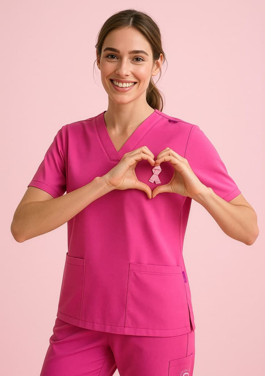 A woman in a bright pink V-neck scrub top makes a heart shape with her hands. The top has a logo.