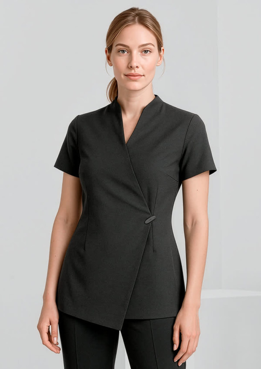 A woman is wearing a black spa tunic with a wrap design and short sleeves. The tunic features a simple button detail.