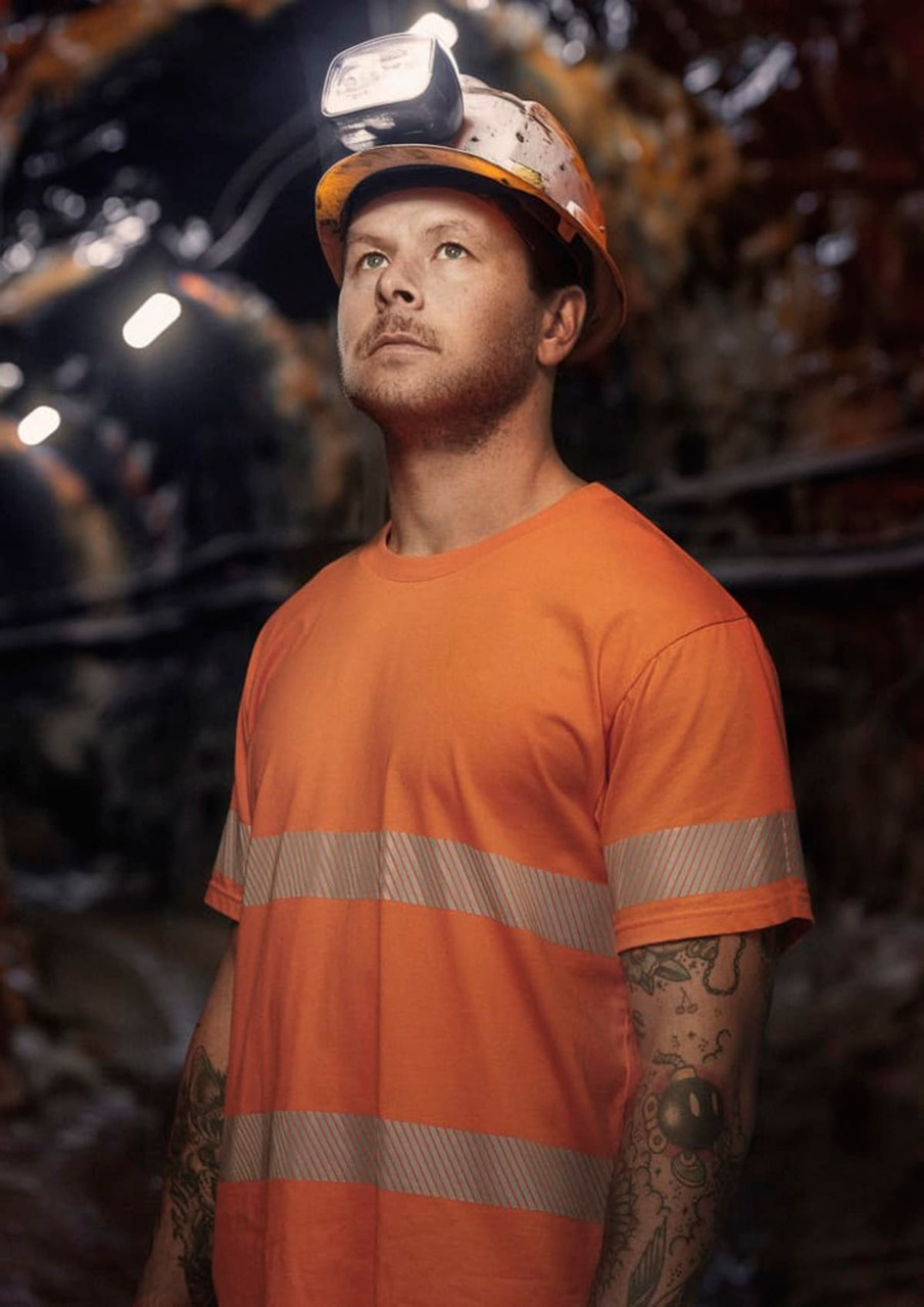 A man wearing an orange hi-vis segmented tape cotton tee shirt and a safety helmet with headlamp, standing in a dimly lit area.