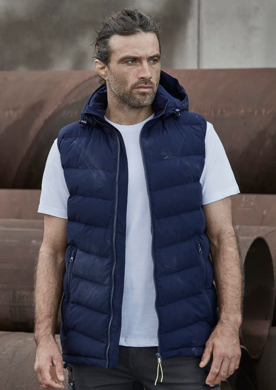 A man wearing a navy blue unisex hooded puffer vest over a white t-shirt, standing in an industrial setting.