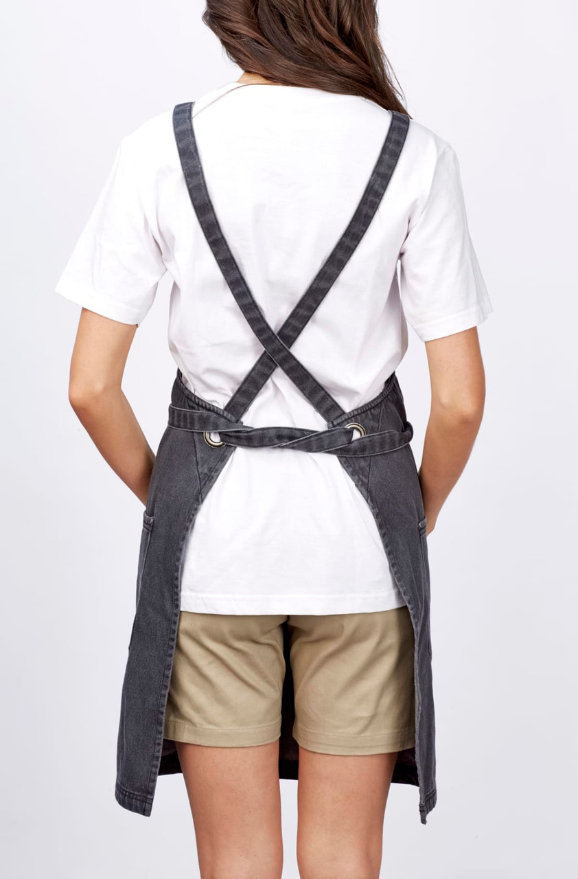 A grey bib apron worn over a white t-shirt, featuring crossed back straps and a casual fit.