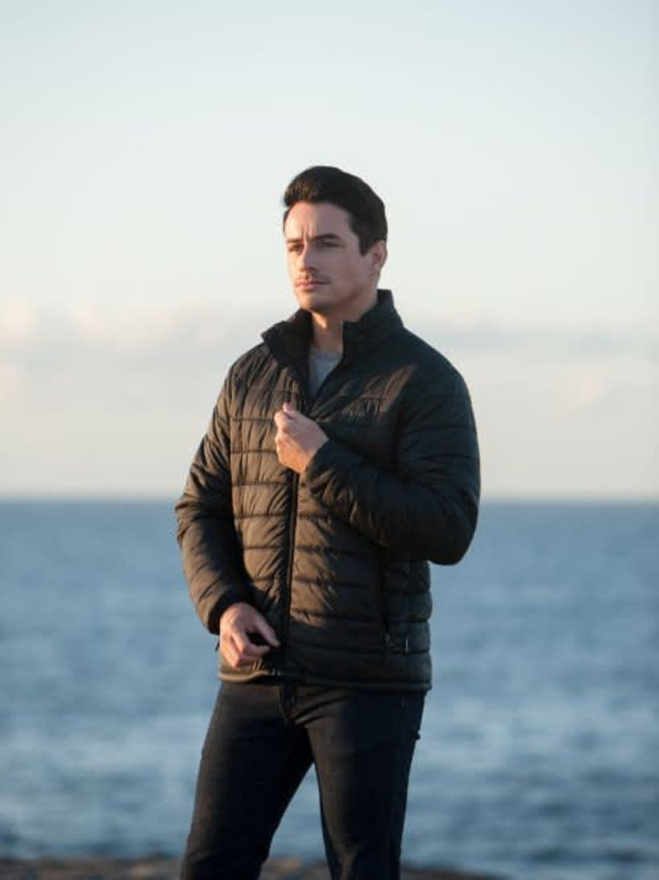 A black puffer jacket for men, featuring a zip-up front and a logo, worn by a model against a coastal backdrop.