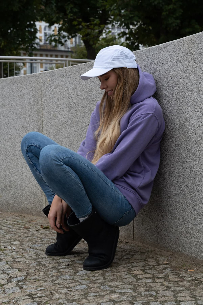 A woman wearing a white cap sits on a grey stone surface, dressed in a purple hoodie and blue jeans.