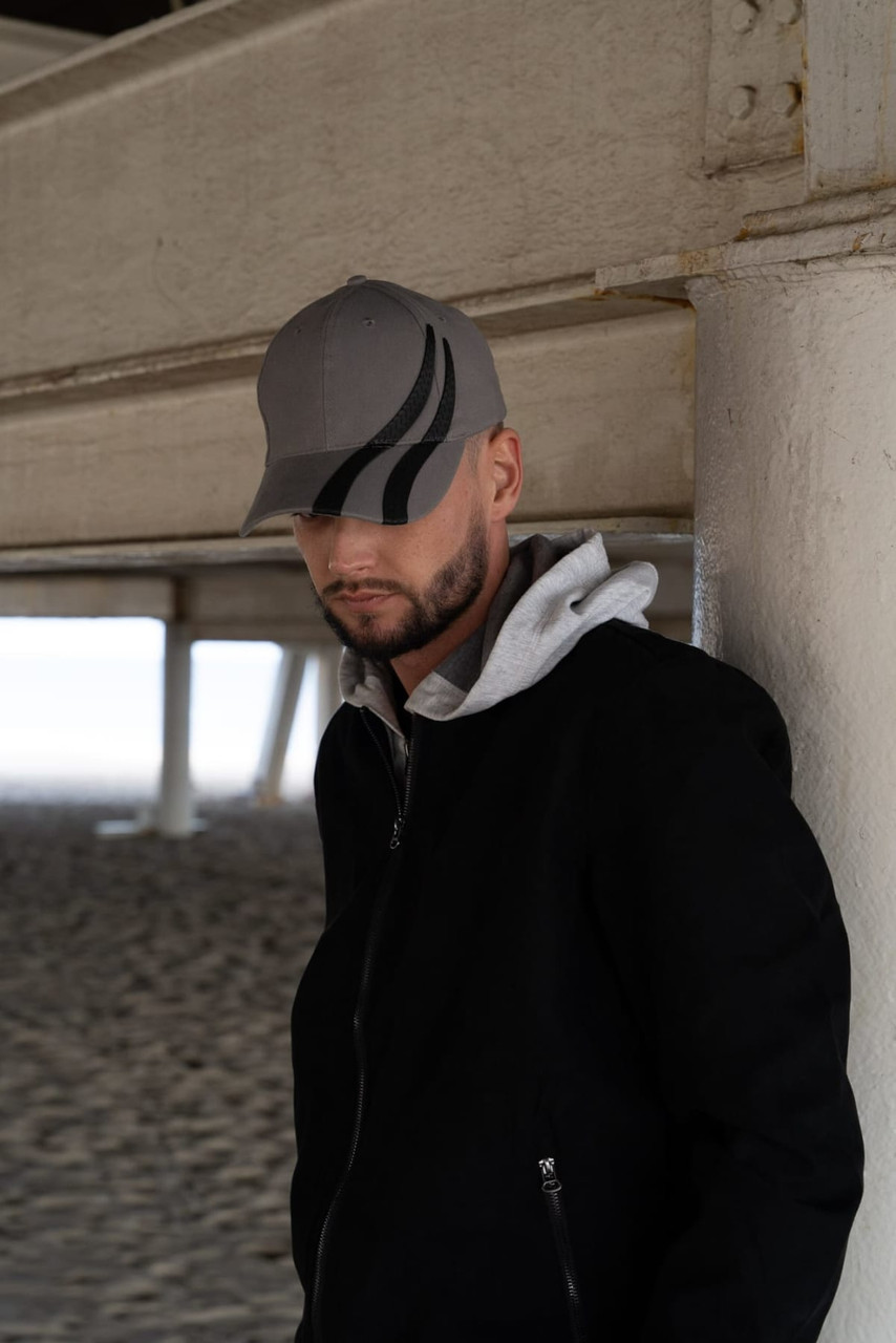 A grey cap with black tyre track designs, worn by a person in a black jacket and grey hoodie under a structure.