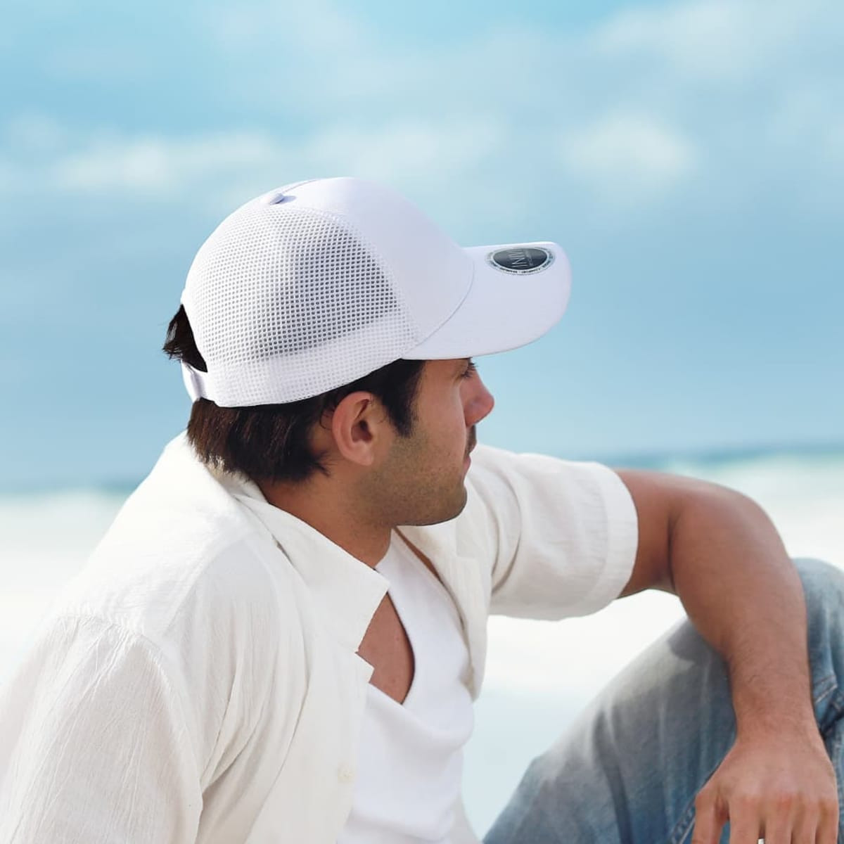 A white mesh cap is worn by a man sitting on a beach, with blue skies and ocean in the background.
