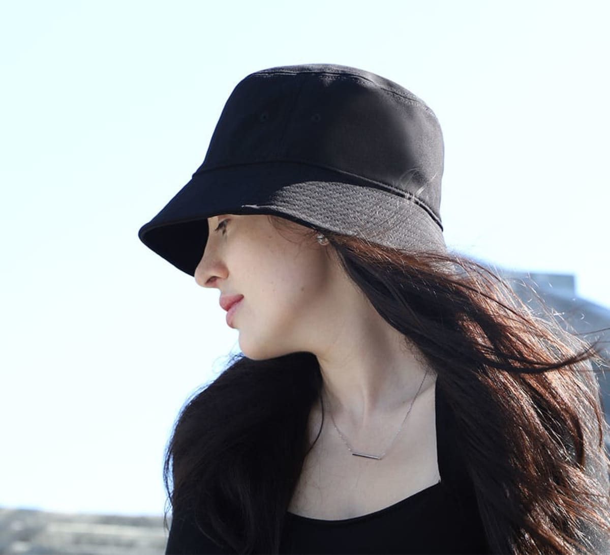 A woman wearing a black bucket hat with a wide brim, set against a bright outdoor background.