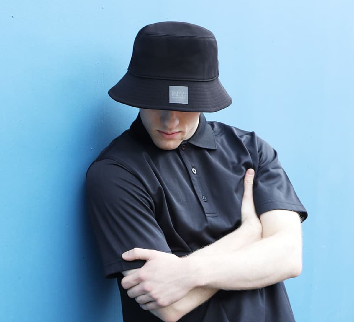 A black bucket hat with a logo, worn by a person in a navy polo shirt against a blue background.