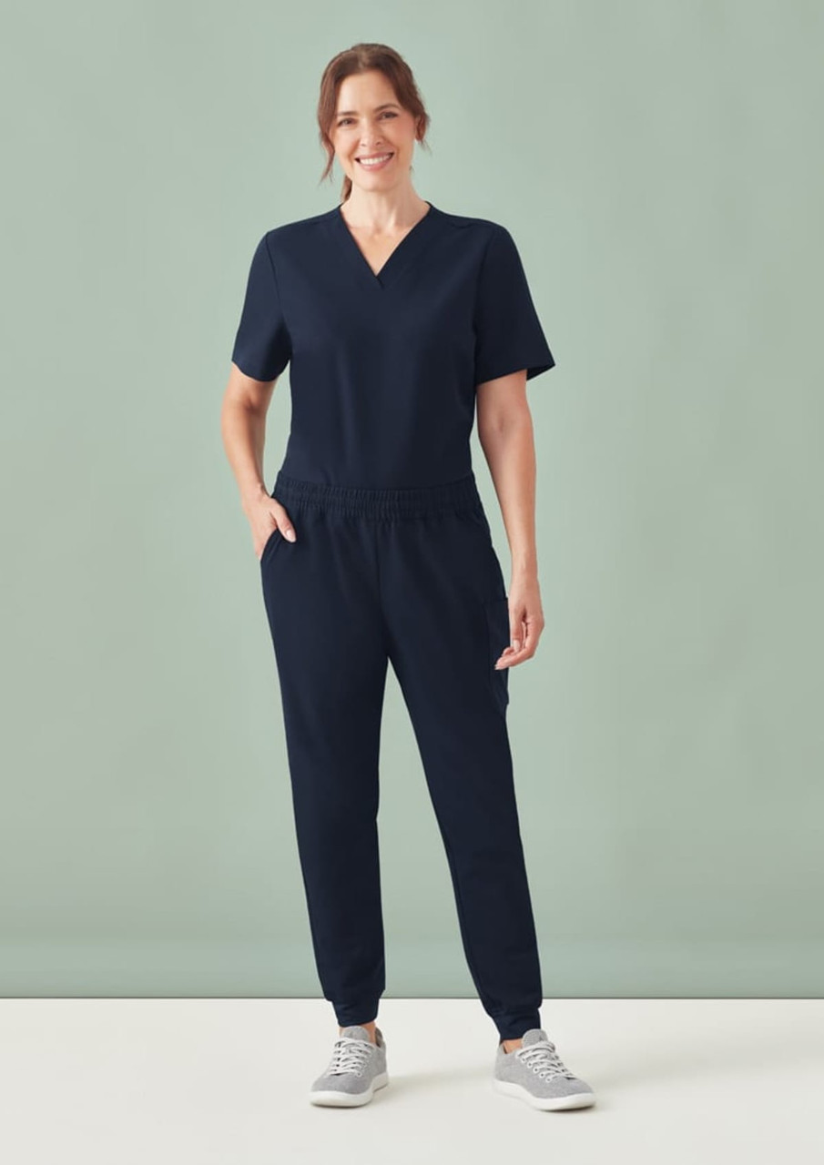 A woman stands wearing navy jogger scrub pants and a matching short-sleeve top, with a casual and comfortable look.