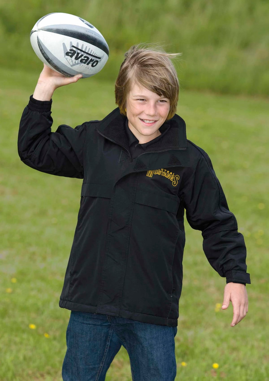 A youth wearing a black rain jacket, holding a rugby ball, standing on grass outdoors. The jacket has a logo.