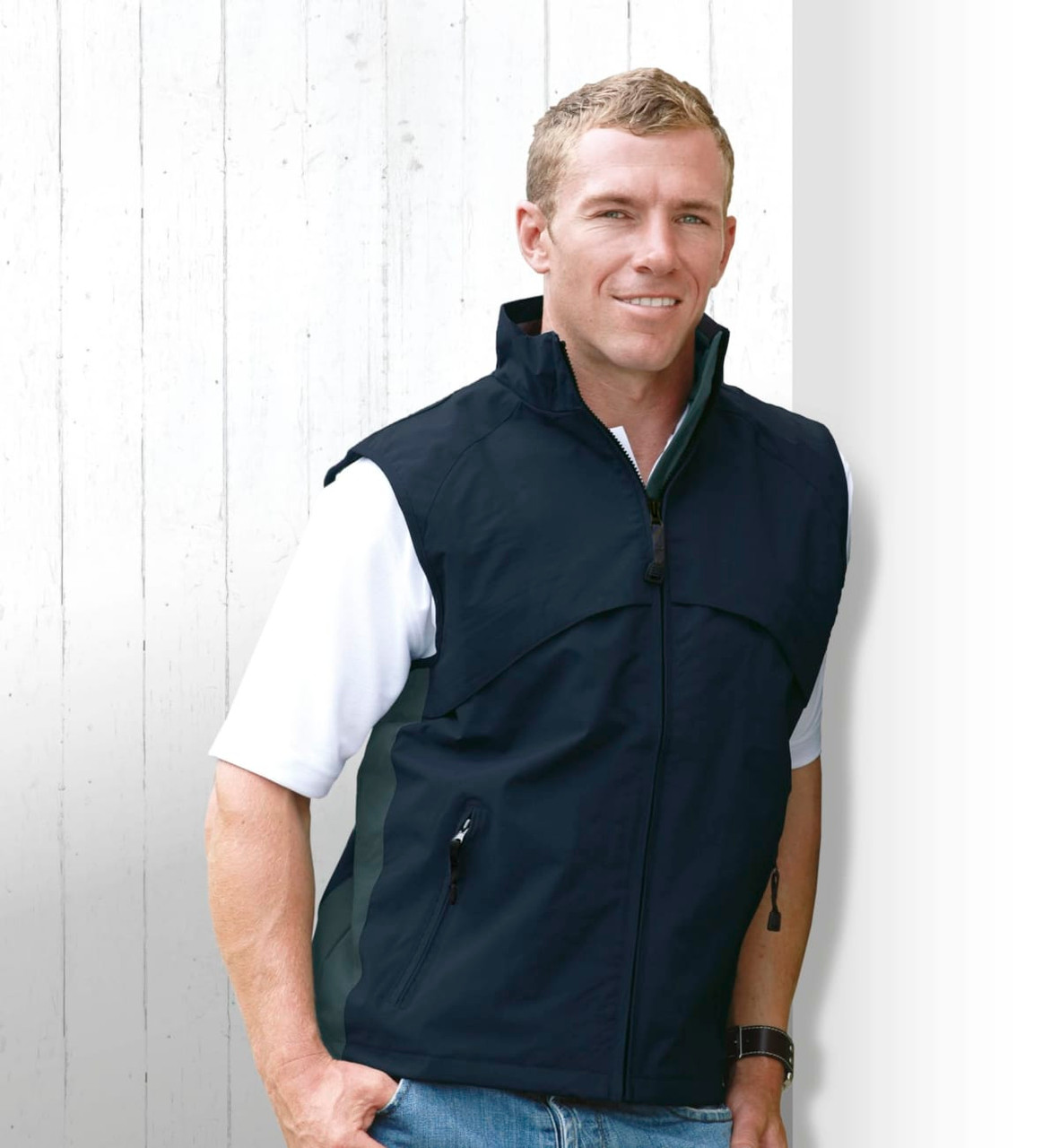 A man wearing a black vest, standing against a white wooden background. The vest features a zip and side pockets.