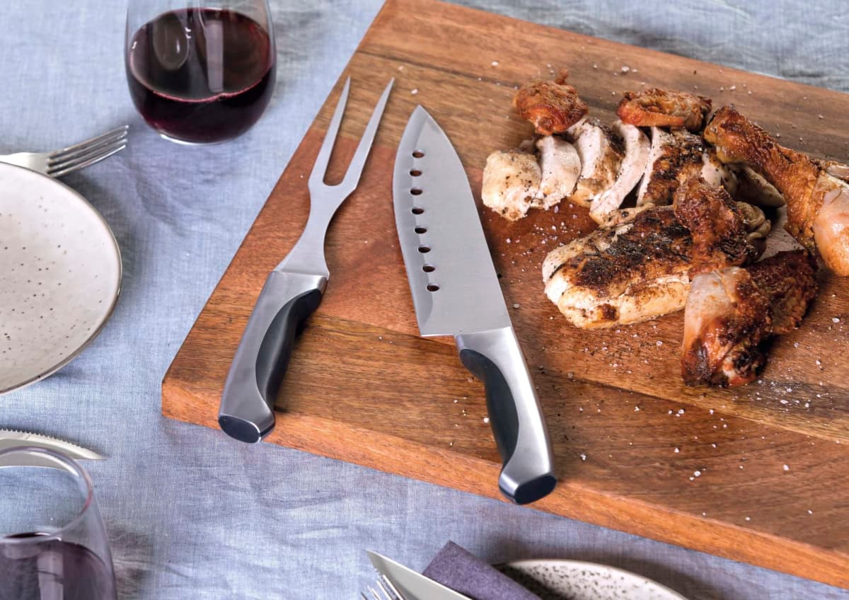 A carving set with a knife and fork on a wooden cutting board, featuring cooked meat. The utensils have black handles.