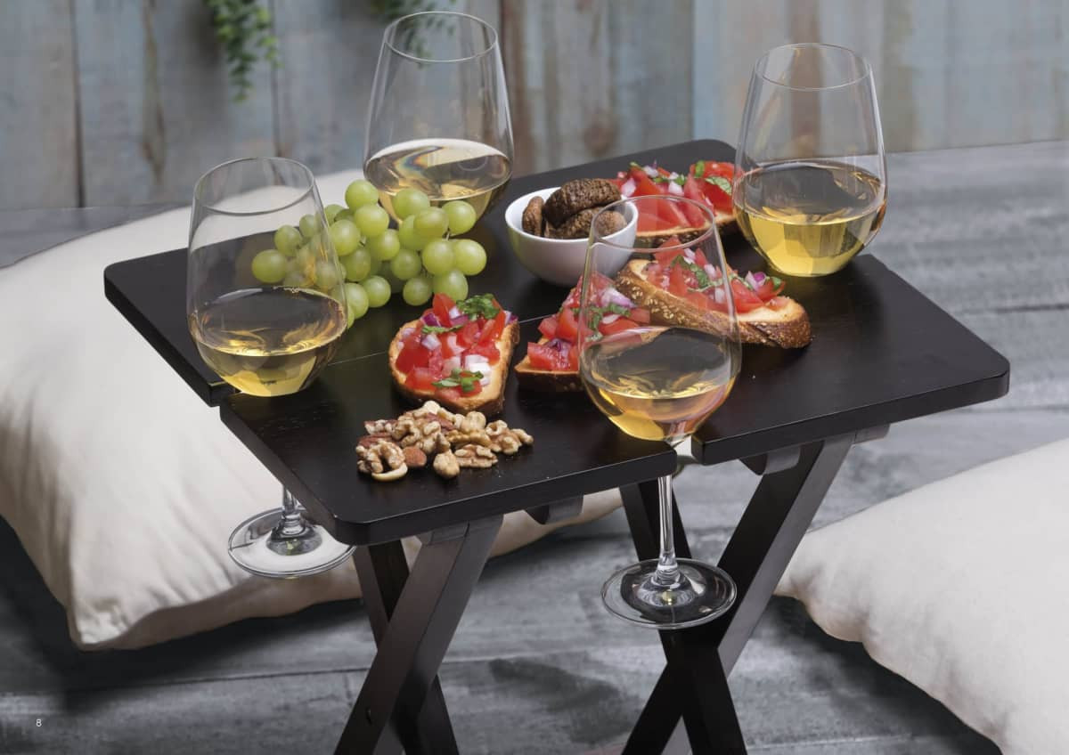 A black table holds glasses of white wine, food platters, grapes, and snacks, creating a leisure outdoor setting.